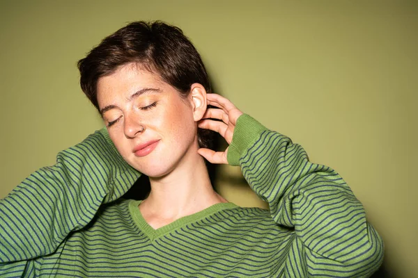 Pleased woman in stylish striped jumper holding hands behind neck and smiling with closed eyes on green background — Stock Photo