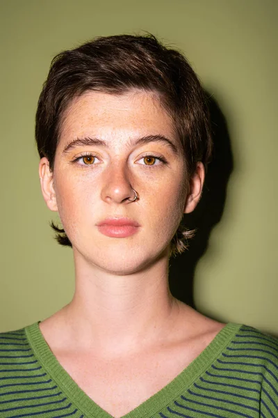Porträt einer brünetten Frau mit Sommersprossen und Nasenpiercing, die in die Kamera auf grünem Hintergrund blickt — Stockfoto