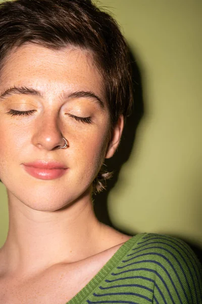 Retrato de mulher sonhadora com sardas e nariz piercing sorrindo com olhos fechados no fundo verde — Fotografia de Stock