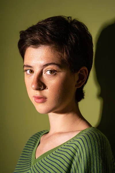 Porträt einer jungen sommersprossigen Frau mit kurzen brünetten Haaren und Nasenpiercing, die auf grünem Hintergrund mit Schatten in die Kamera blickt — Stockfoto