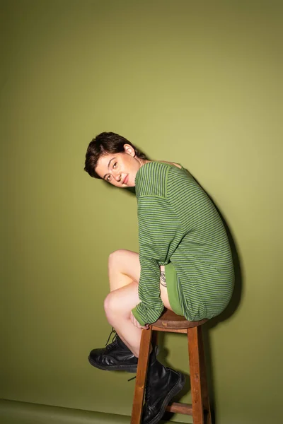 Comprimento total de mulher tatuada sorridente em pulôver listrado e botas pretas posando em banquinho de madeira em fundo verde — Fotografia de Stock
