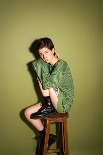 Volle Länge der trendigen Frau in gestreiftem Pullover und schwarzen Lederstiefeln sitzt auf einem Holzhocker auf grünem Hintergrund mit Schatten — Stockfoto