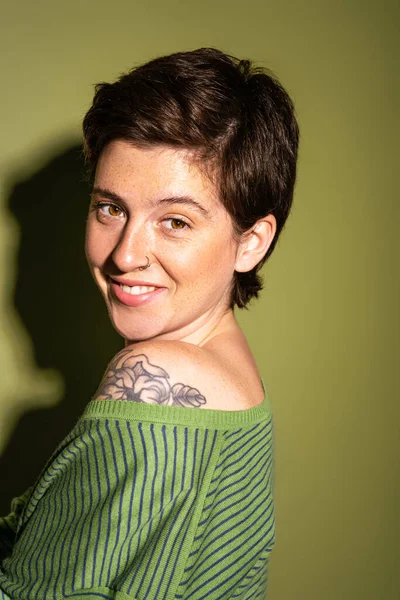 Портрет радостной женщины с веснушками и татуировкой, улыбающейся в камеру на зеленом фоне с тенью — стоковое фото