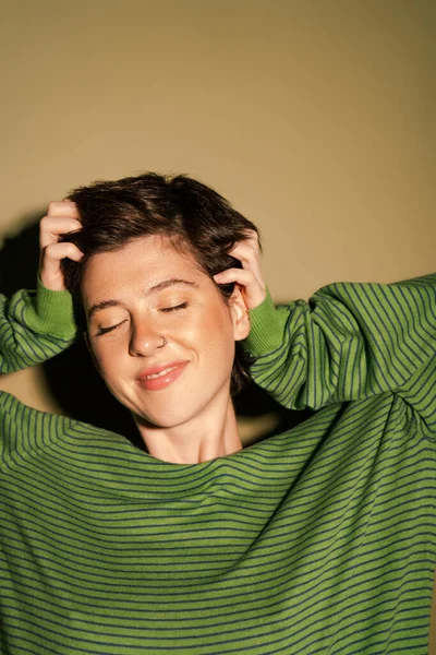 Довольная женщина в полосатом пуловере трогает короткие брюнетки и улыбается с закрытыми глазами на зеленом фоне — стоковое фото