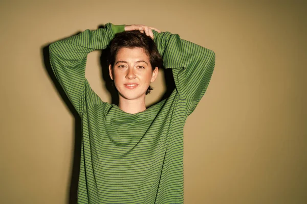 Happy brunette woman in trendy striped jumper posing with hands above head and smiling at camera on green background — Stock Photo