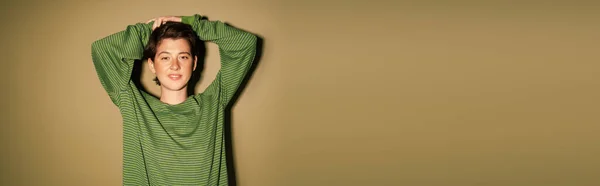 Веселая женщина в полосатом свитере позирует с руками над головой и улыбается в камеру на зеленом фоне, баннер — стоковое фото
