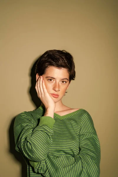 Displeased brunette woman in striped jumper looking at camera while holding hand near face on green background — Stock Photo