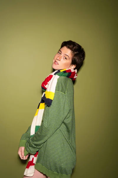 Молодая и позитивная женщина в модном свитере и полосатом разноцветном шарфе, смотрящая на камеру на зеленом фоне — стоковое фото