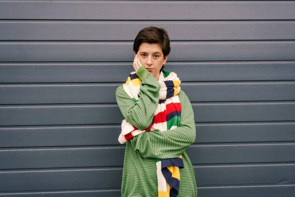 Скептична жінка в зеленому пуловері і смугастий різнокольоровий шарф позує рукою біля обличчя на сірій стіні на вулиці — стокове фото