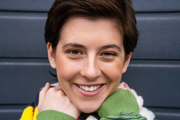 Portrait of joyful brunette woman with freckles and nose piercing smiling at camera on street in spring — Stock Photo