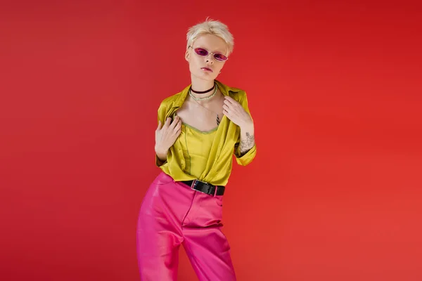 Татуированная модель альбиноса в стильном наряде и модные солнцезащитные очки, позирующие на карминовом розовом фоне — стоковое фото