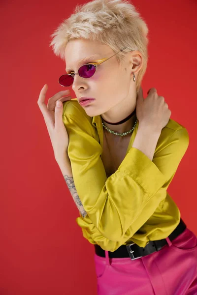 Модель альбиноса с татуированной рукой, позирующей в стильном наряде и модных солнцезащитных очках на кармине розового цвета — стоковое фото