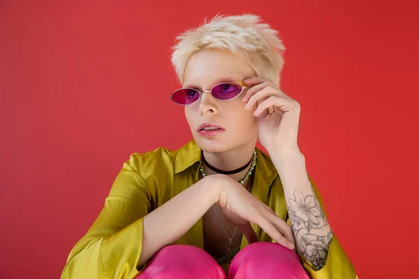 Modelo albino con tatuaje posando en blusa elegante y ajustando gafas de sol de moda sobre fondo rosa carmín - foto de stock