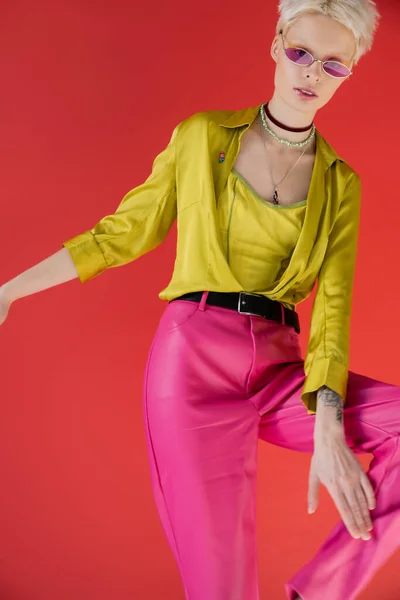 Альбіно модель з татуйованою рукою позує в стильній блузці та рожевих сонцезахисних окулярах на кармінному фоні — стокове фото