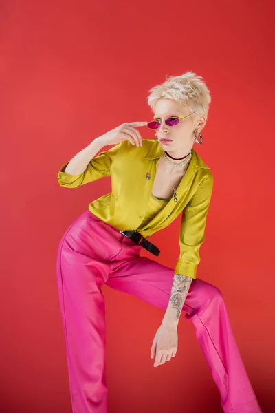 Albino model with tattoo posing in stylish blouse and adjusting trendy sunglasses on carmine pink — Stock Photo
