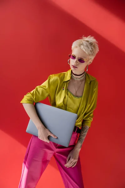 Blonde Albino-Frau mit Tätowierung posiert in trendiger Sonnenbrille und hält Laptop auf karminrosa — Stockfoto