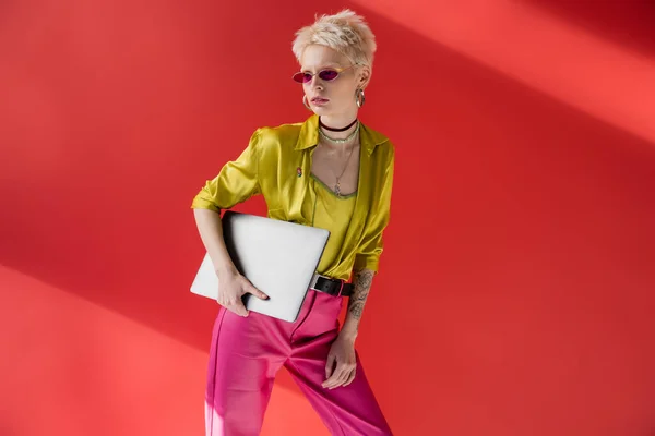 Stylische Albino-Freelancer mit Tätowierung posiert in trendiger Sonnenbrille und hält Laptop auf karminrosa Hintergrund — Stockfoto