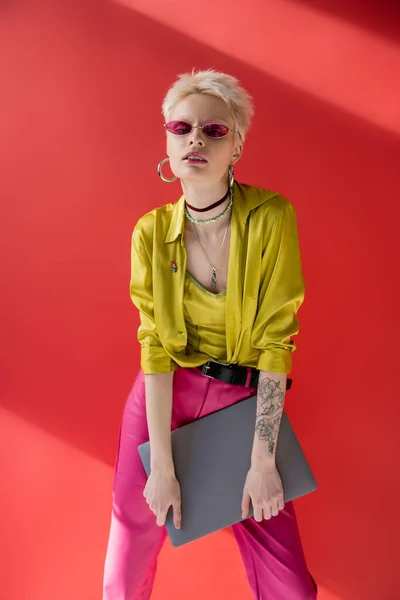 Rubia albino freelancer con tatuaje posando en gafas de sol de moda y sosteniendo portátil en carmine rosa - foto de stock
