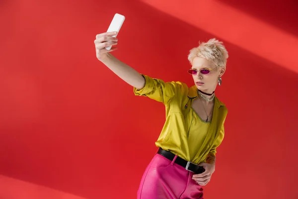 Tattooed albino woman in stylish sunglasses taking selfie on mobile phone on carmine pink background — Stock Photo