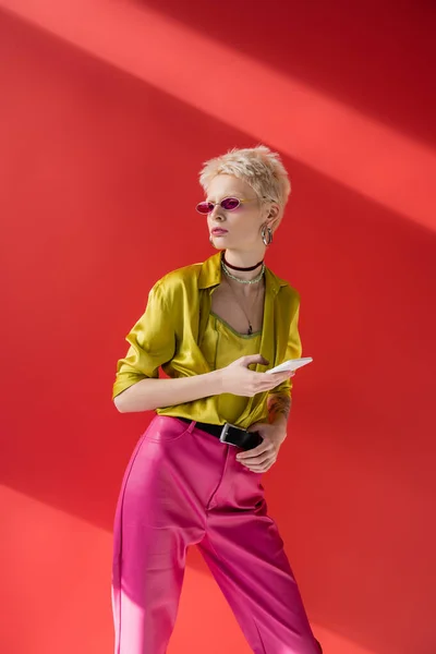 Tätowierte Albino-Frau in trendiger Sonnenbrille mit Smartphone auf karminrosa — Stockfoto