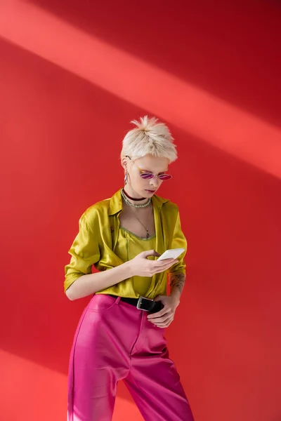 Tätowierte Albino-Frau mit trendiger Sonnenbrille textet auf Smartphone auf karminrosa — Stockfoto