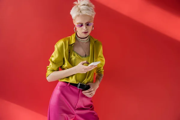 Татуированная альбиноса женщина в модных солнцезащитных очках сообщения на смартфоне на кармине розовый фон — стоковое фото