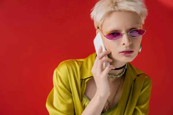Albino woman in trendy sunglasses talking on smartphone on carmine pink background — Stock Photo