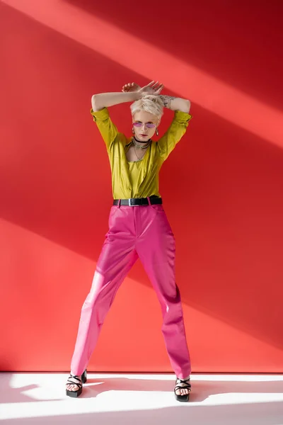 Полная длина блондинка с татуированной рукой, позируя в стильных сандалиях на каблуках и модных солнцезащитных очках на кармине розовый — стоковое фото