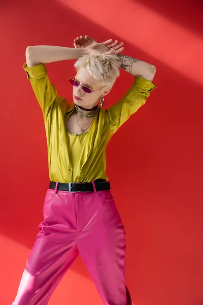 Блондинка с татуированной рукой, позирующая в стильной блузке и модных солнцезащитных очках на кармине розовый — стоковое фото