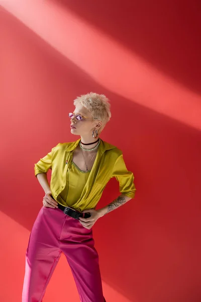 Блондинка модель з татуюванням на руці позує в стильній блузці і модних сонцезахисних окулярах на карміновому рожевому — стокове фото