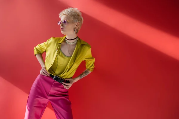 Молодая модель альбиноса с татуировкой на руке, позирующая в стильной блузке и модных солнцезащитных очках на розовом фоне — стоковое фото
