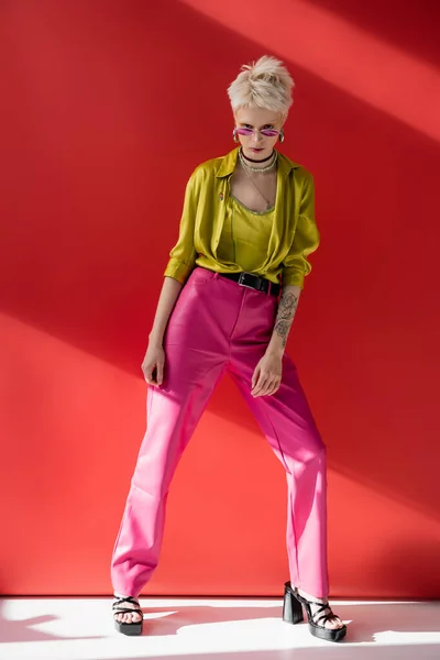 Полная длина молодой модели альбиноса с татуировкой на руке, позирующей в стильных сандалиях на каблуках и модных солнцезащитных очках на кармине розовый — стоковое фото