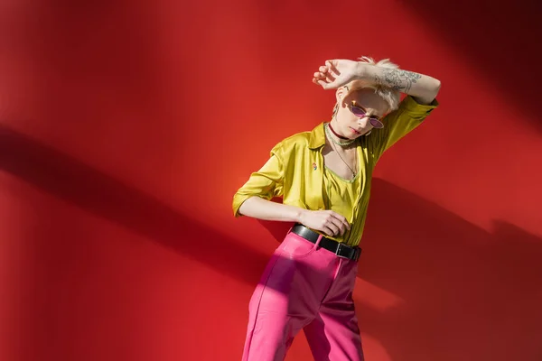 Солнечный свет на лице модели альбиноса с татуировкой, позирующей в стильных солнцезащитных очках на розовом фоне — стоковое фото