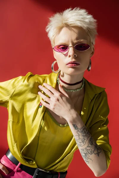 Luz del sol en la cara de la mujer albina con tatuaje en la mano posando en gafas de sol elegantes en rosa carmín - foto de stock