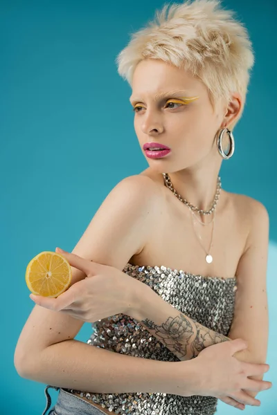 Tattooed blonde albino woman with bare shoulders holding lemon half on blue — Stock Photo