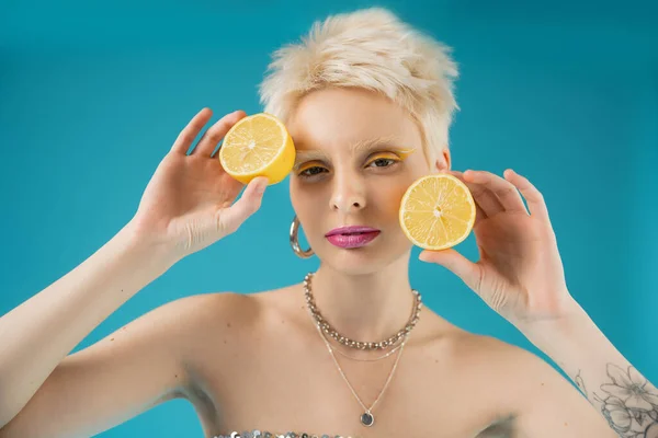 Blonde albino model with tattoo on hand holding sour lemon halves on blue background — Stock Photo