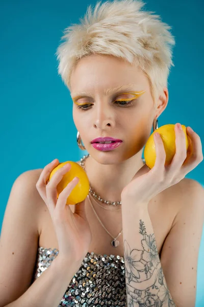 Татуированная модель альбиноса с модным макияжем и обнаженными плечами, позирующими с лимонами на синем — стоковое фото