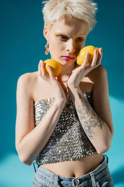 Mujer albina tatuada con maquillaje de moda y cabello rubio posando con limones maduros en azul - foto de stock