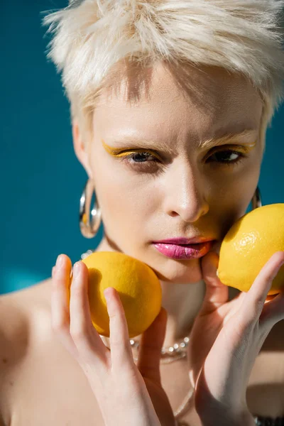 Портрет альбінової жінки з модним макіяжем і світлим волоссям, позує зі стиглими лимонами на синьому — стокове фото