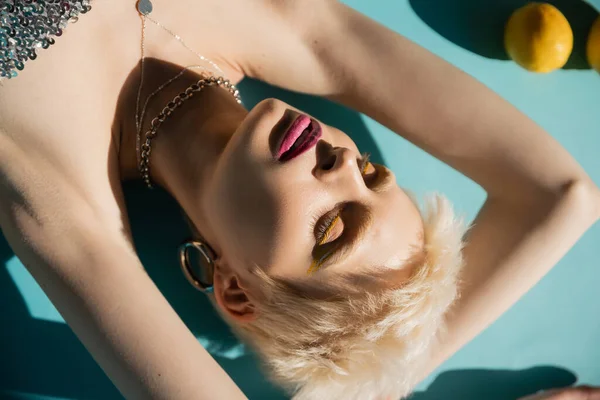 Vista dall'alto della donna albina con gli occhi chiusi e trucco alla moda in posa vicino a limoni maturi su blu — Foto stock