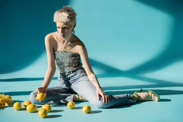 Full length view of albino woman in shiny top with sequins sitting near ripe lemons on blue background — Stock Photo
