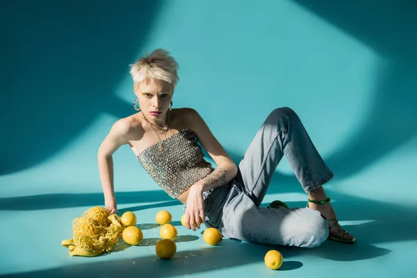 Ganzkörperansicht tätowierter Frau in glänzendem Top mit Pailletten, die in der Nähe reifer Zitronen auf blauem Hintergrund sitzt — Stockfoto