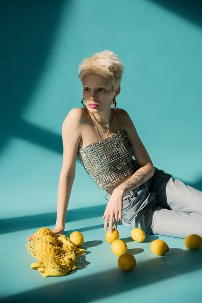 Tattooed albino model in shiny top with sequins and jeans sitting near ripe lemons on blue — Stock Photo
