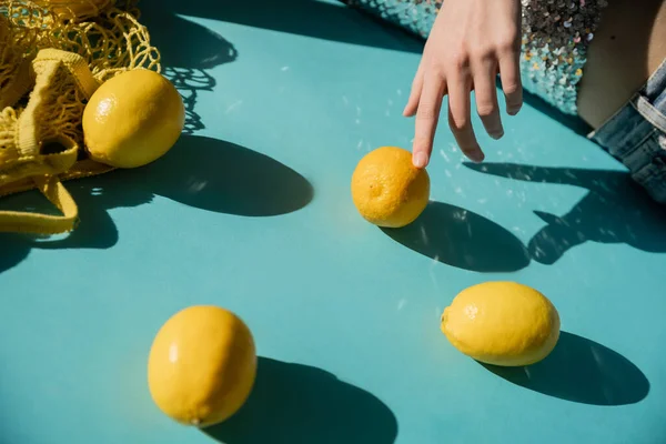 Обрезанный вид женщины в блестящем топе с блестками, позирующими рядом с мешком для струн и спелыми лимонами на голубом — стоковое фото