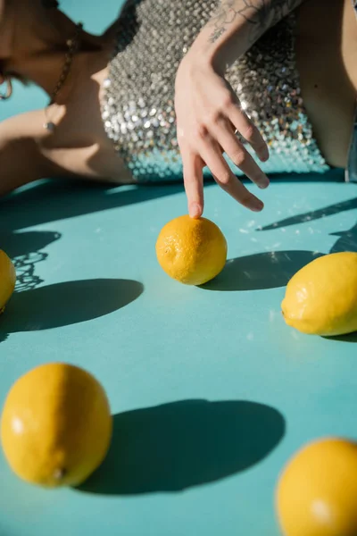 Teilansicht einer tätowierten Frau in glänzendem Top mit Pailletten, die in der Nähe von String Bag und reifen Zitronen auf Blau posieren — Stockfoto