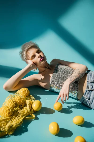 Tattooed albino model in shiny top with sequins and jeans posing near ripe lemons on blue — Stock Photo