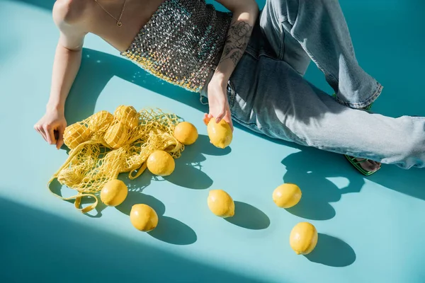 High angle view of tattooed woman in shiny top with sequins and jeans sitting near string bag and ripe lemons on blue — Stock Photo