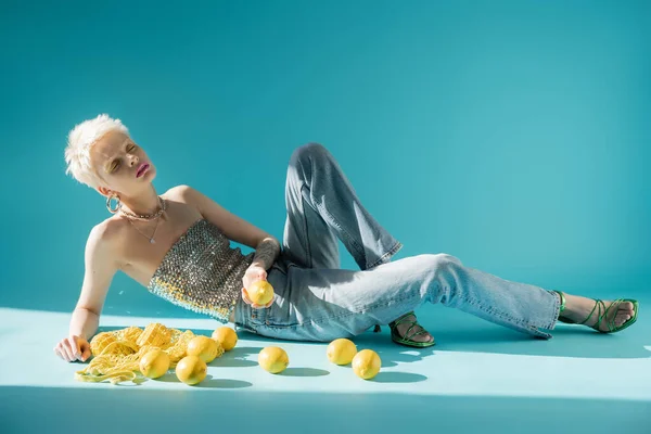 Vista a figura intera di donna albina tatuata in alto con paillettes e jeans in posa vicino a limoni freschi su blu — Foto stock