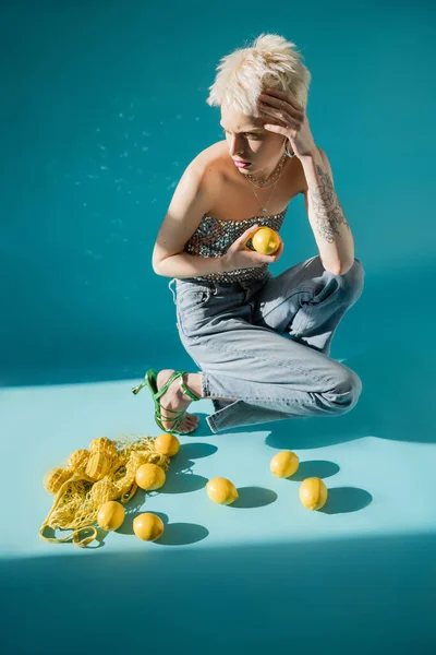Vue pleine longueur de femme albinos tatoué en haut avec des paillettes et jeans en denim posant près de citrons frais sur bleu — Photo de stock