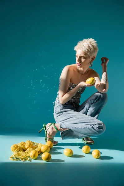 Vista a figura intera della donna albina in alto con paillettes e jeans in denim in posa vicino a limoni freschi su blu — Foto stock